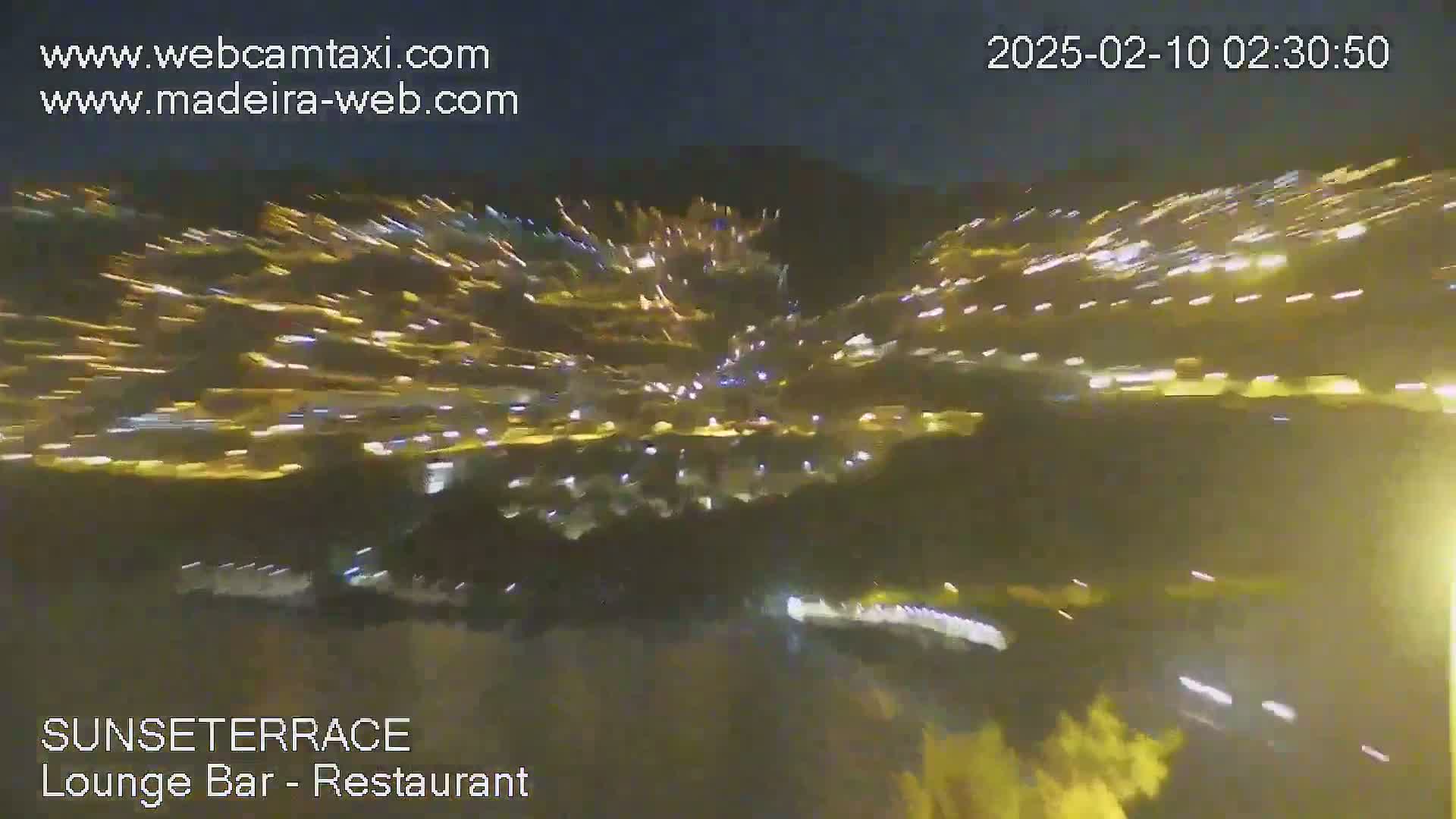 A nighttime long-exposure shot shows streaks of light from vehicles moving along a road below, possibly in a mountainous area.