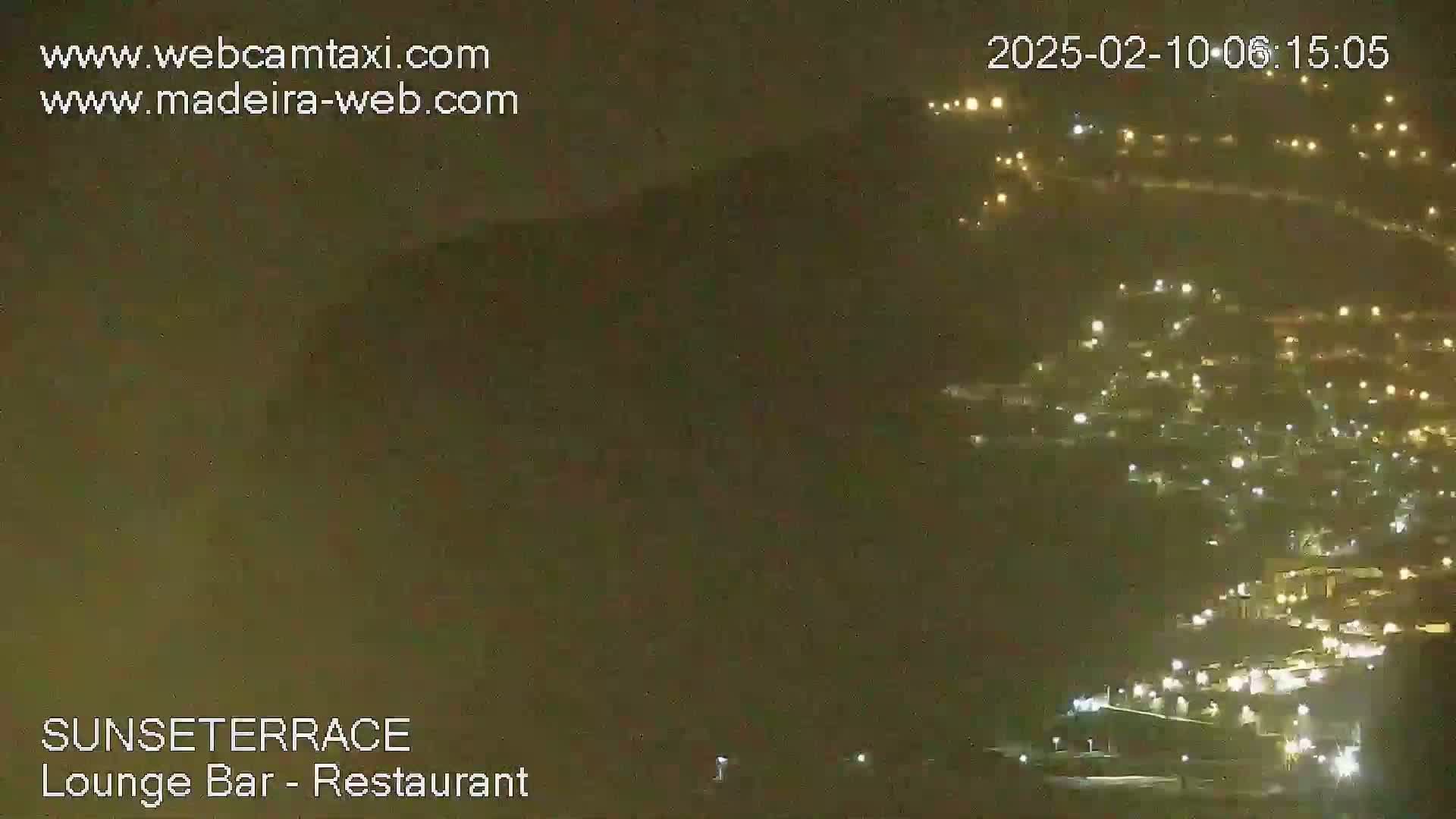 A nighttime long shot shows a hillside town illuminated by many lights under hazy, dark conditions.