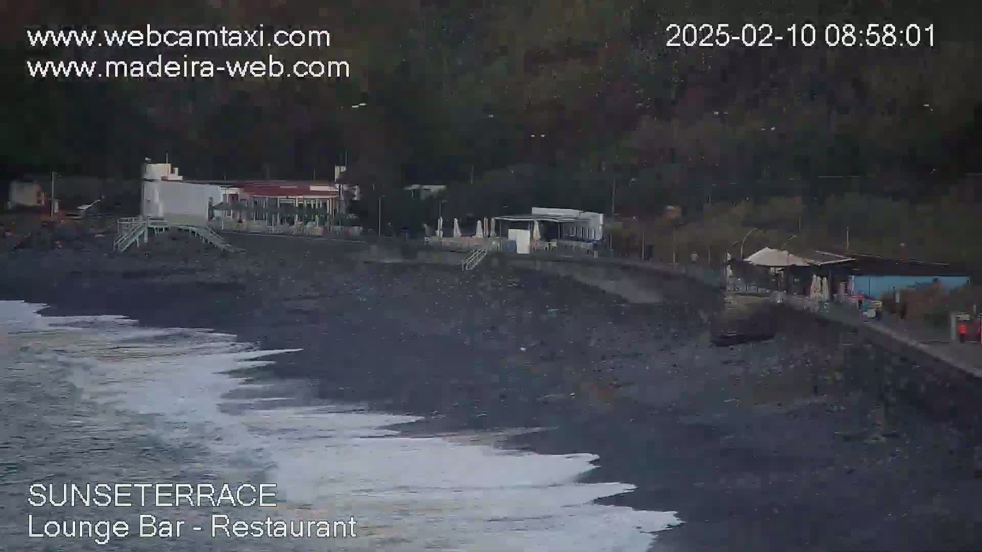 Waves lap a dark-colored beach next to a shoreline restaurant and a hill covered in dark green vegetation under a lightly overcast sky.