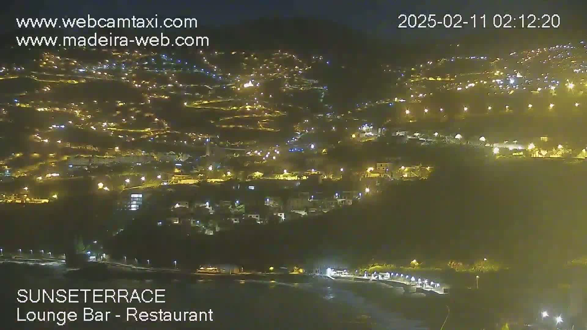 A nighttime, high-angle view of a hillside town illuminated by numerous lights, stretching down to a dark body of water.