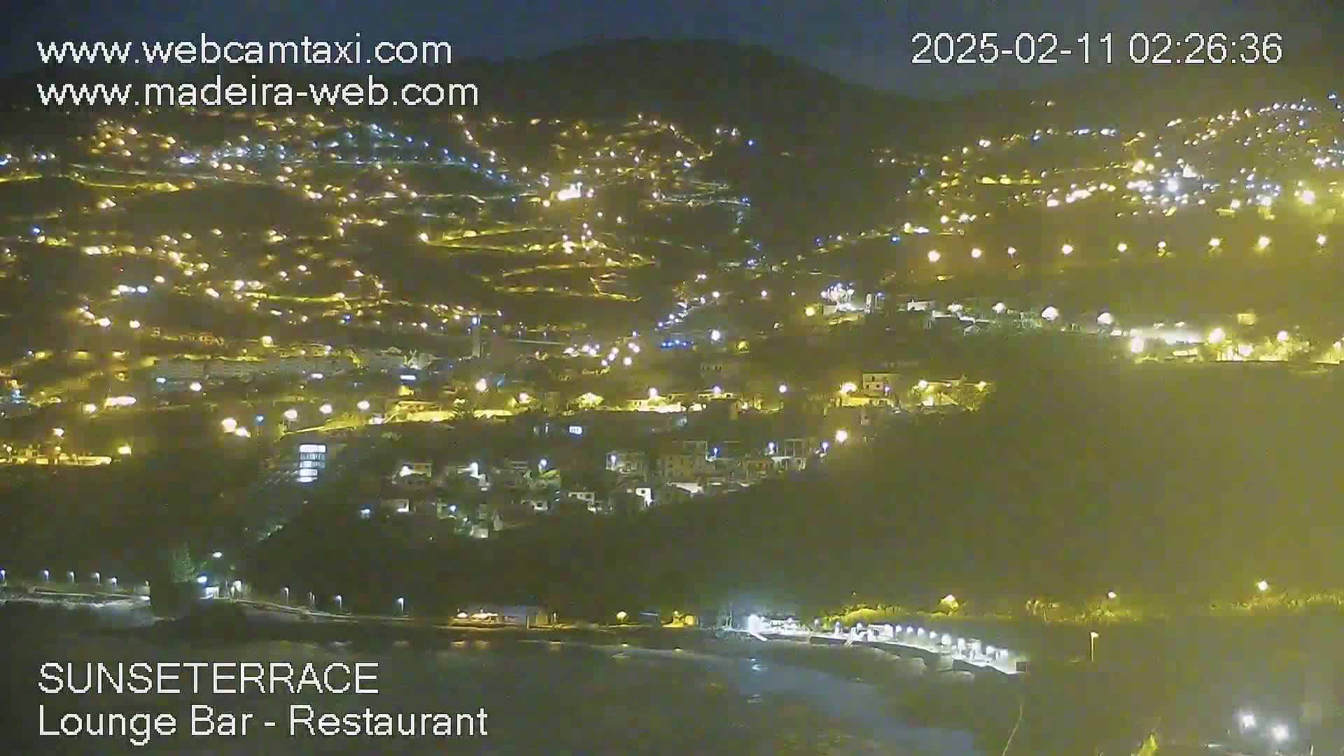 A nighttime, hillside town is illuminated by many small lights, with a dark body of water visible in the foreground.