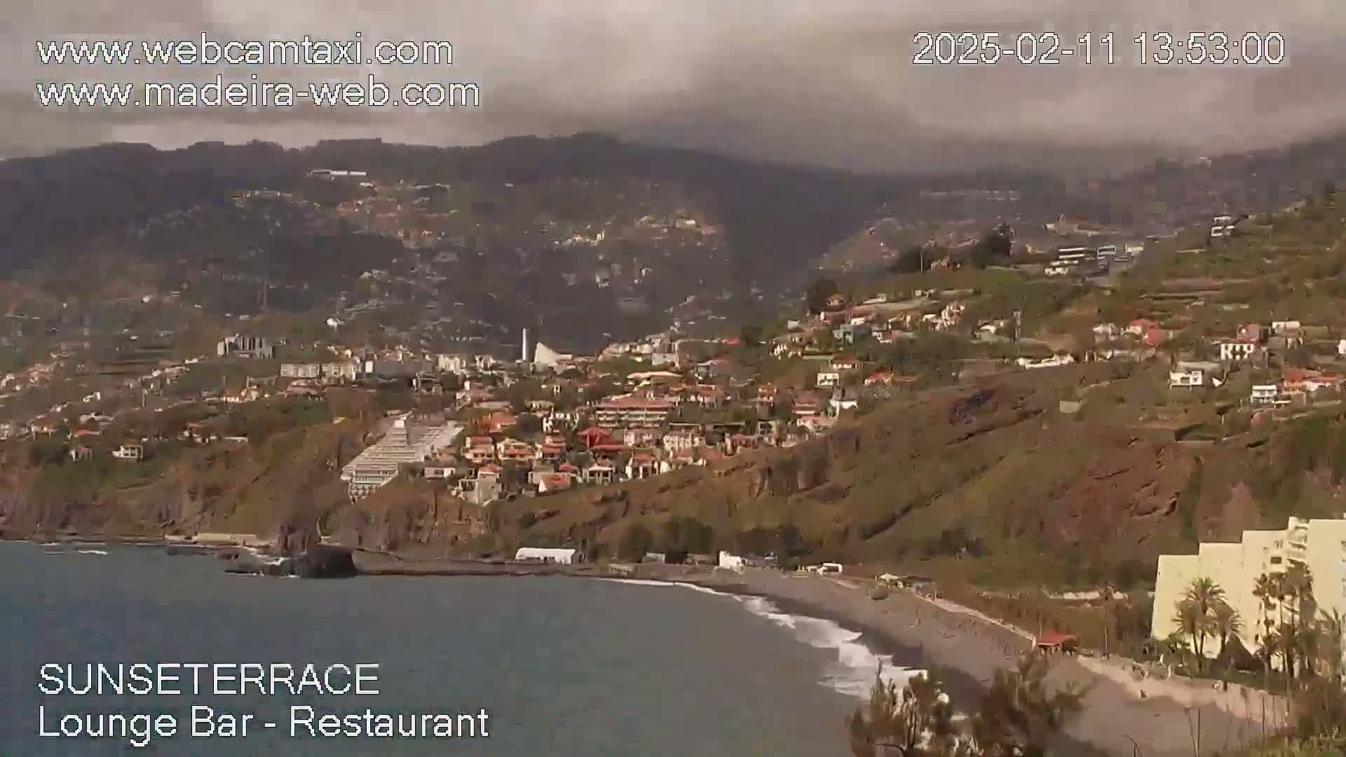 A coastal town nestled on a hillside overlooking a dark-sand beach and ocean, under an overcast sky.