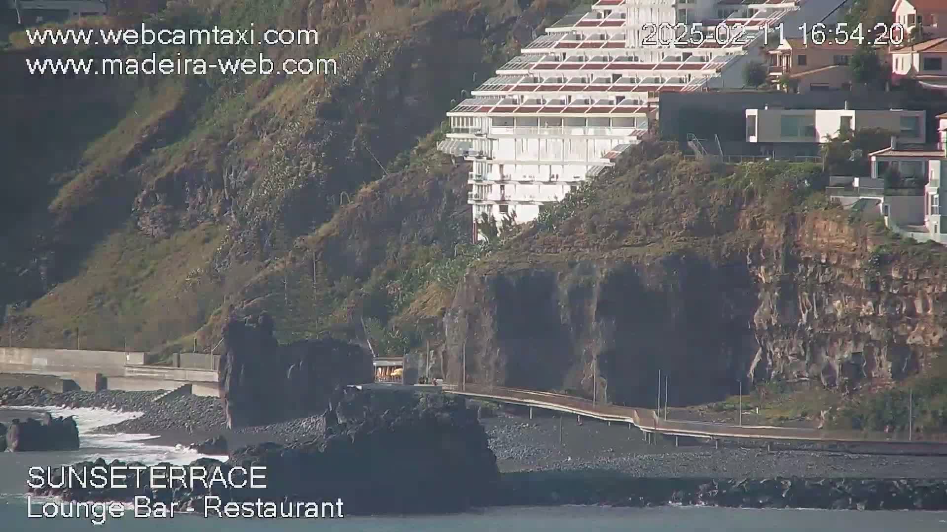 A seaside cliffside with a large white building, a paved walkway, and dark volcanic rock beach is shown on a sunny day.