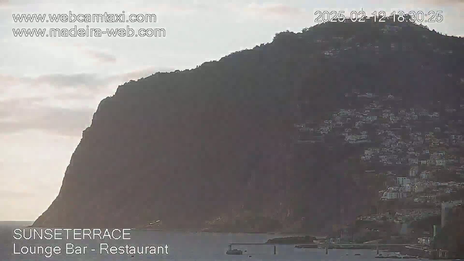A large, dark hill covered in buildings is seen at dusk, overlooking a calm body of water.