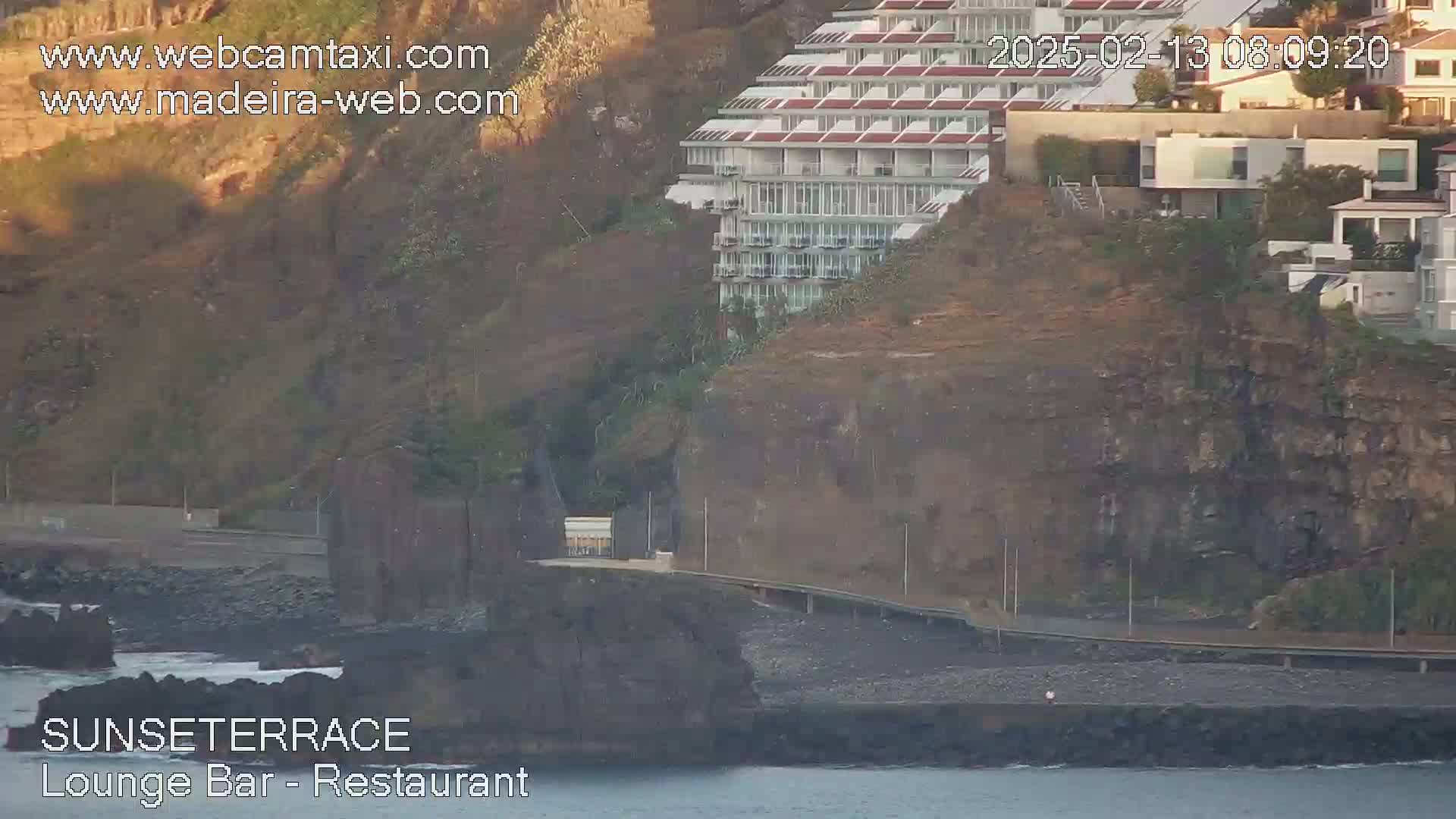 A multi-story building sits atop a rocky cliff overlooking a calm body of water during the daytime.