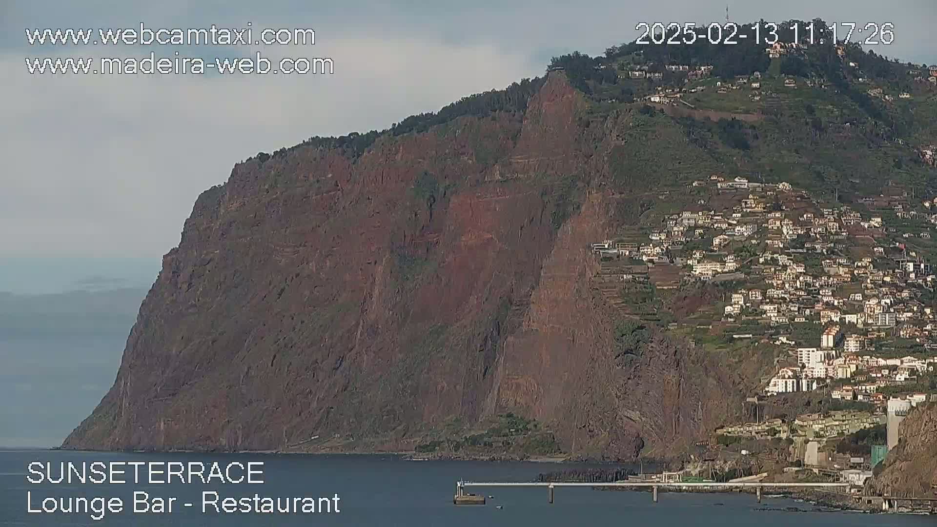 A large, dark-colored cliff overlooks a town and the ocean under a mostly cloudy sky.