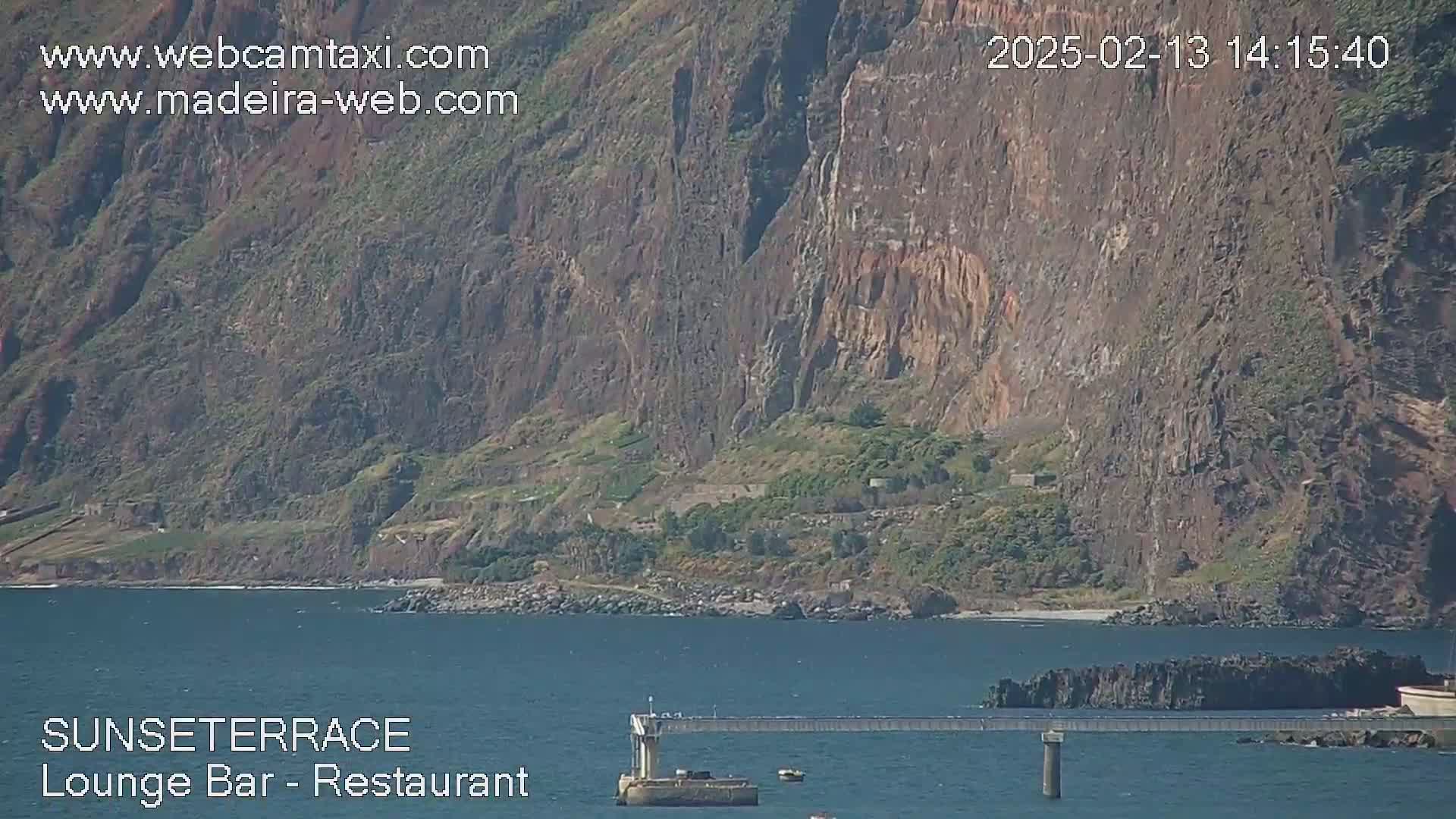 A rocky cliff face overlooks a calm body of water with a pier extending from the shore on a sunny day.