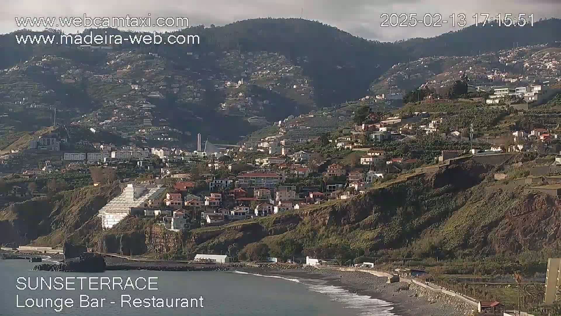 A coastal town nestled on a hillside overlooking a dark-sand beach, under a mostly clear sky.