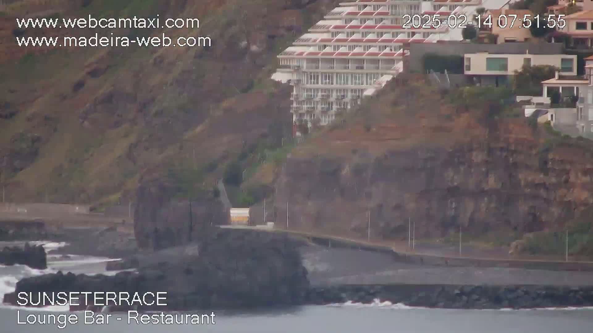 A coastal scene shows a multi-story building perched on a rocky cliff overlooking a dark-colored beach and ocean with small waves.