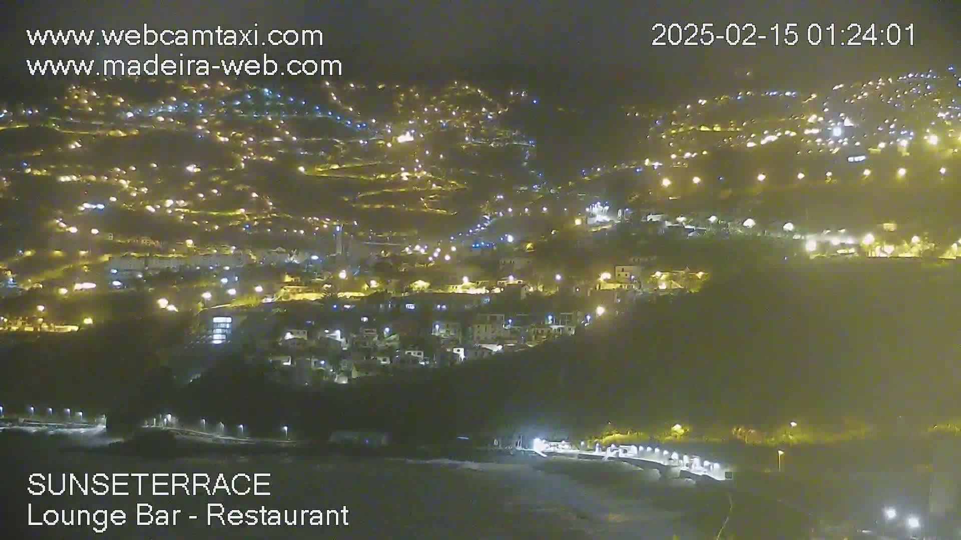 A nighttime, long-range view of a hillside town illuminated by numerous lights, overlooking a dark body of water.