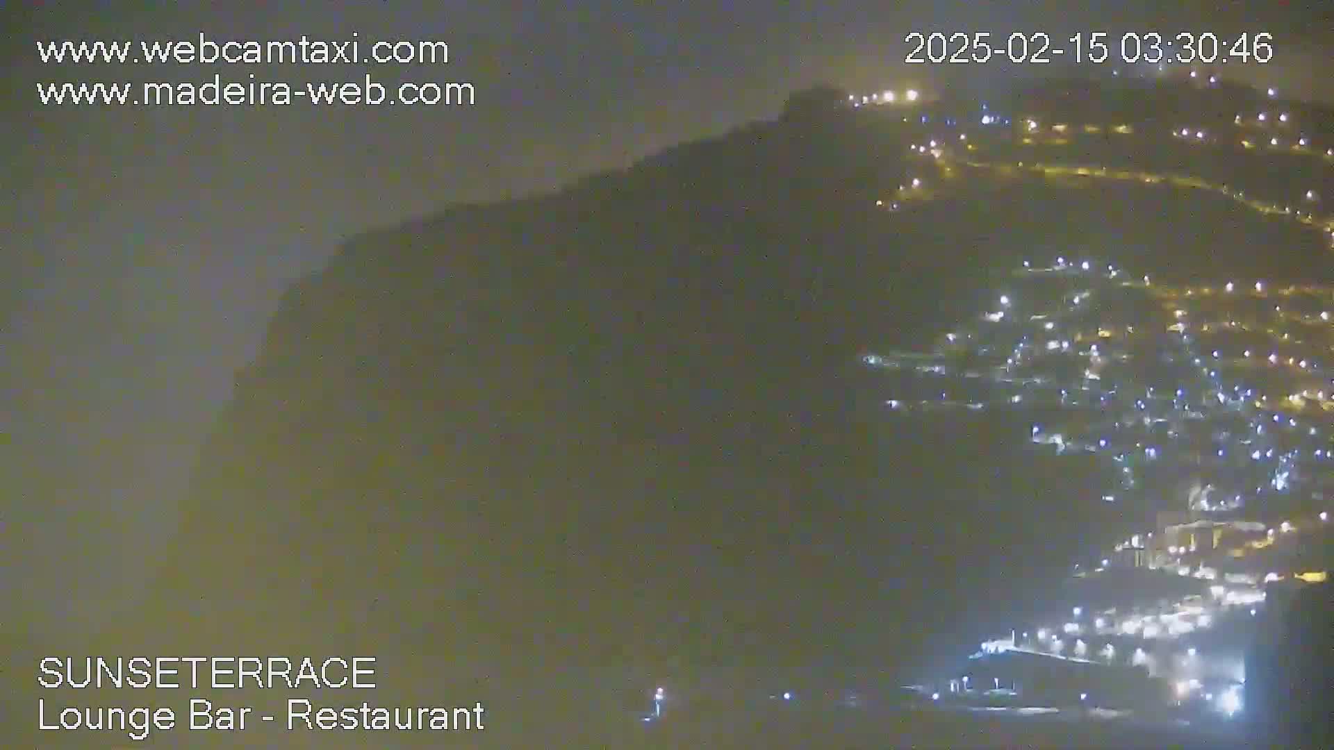 A hazy night scene shows a hillside town illuminated by many lights, with a dark cliff face visible in the background.
