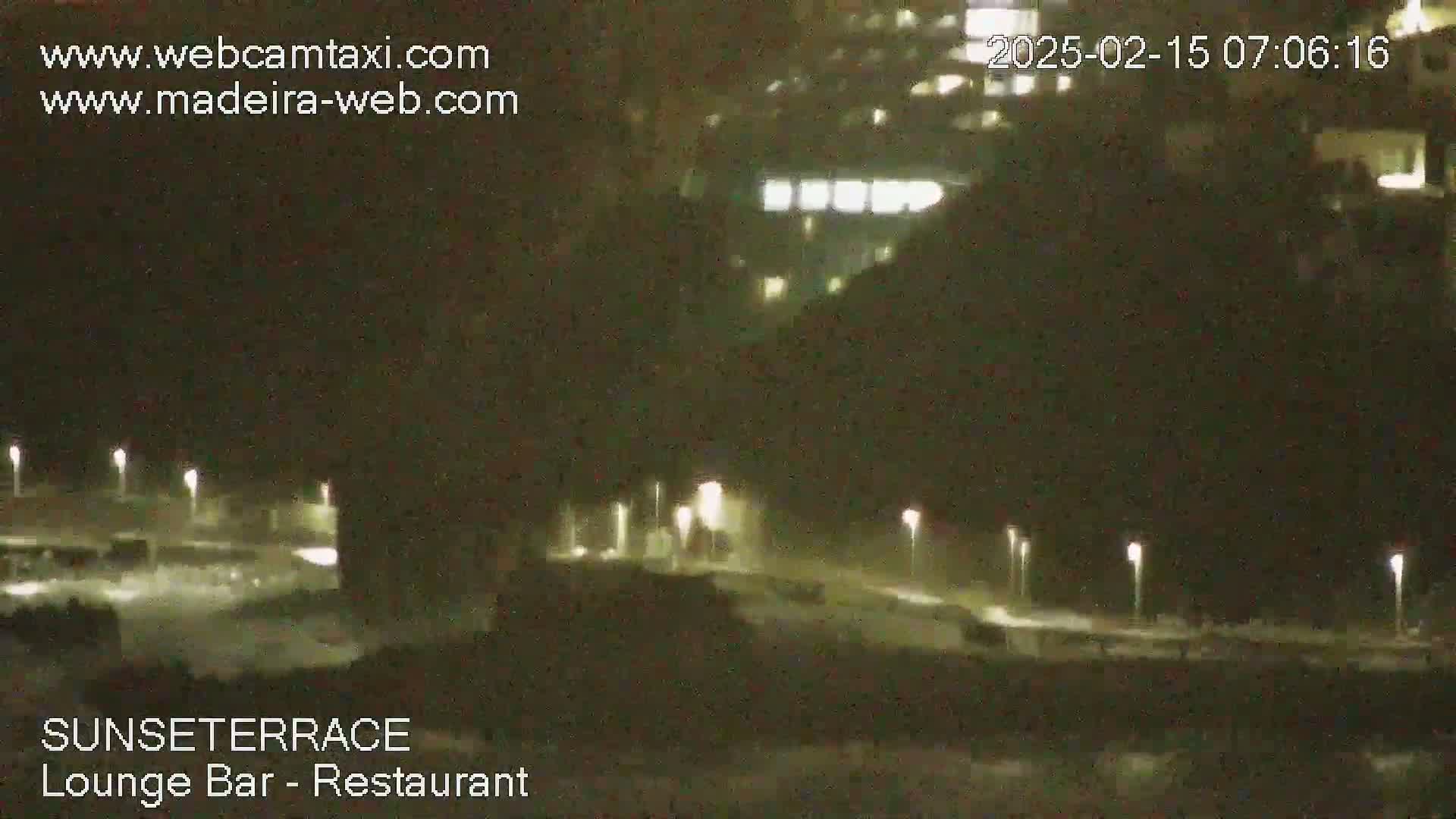 A nighttime, low-light view of an outdoor area with several lit street lamps, a building in the background, and what appears to be a shoreline or waterfront in the foreground.