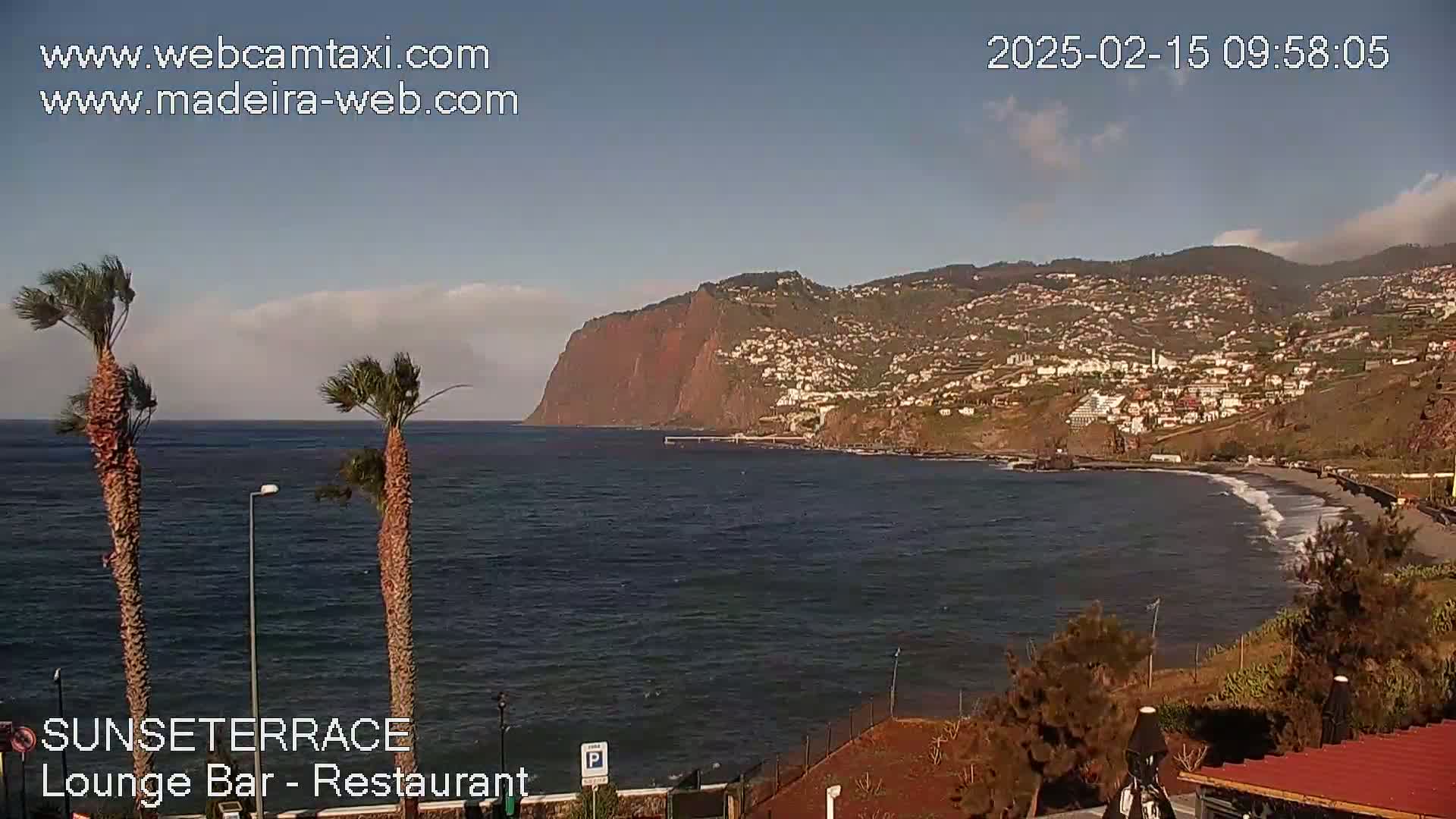 A partly cloudy day reveals a coastal town nestled against a high, reddish cliff overlooking a dark-blue ocean with a small sandy beach.