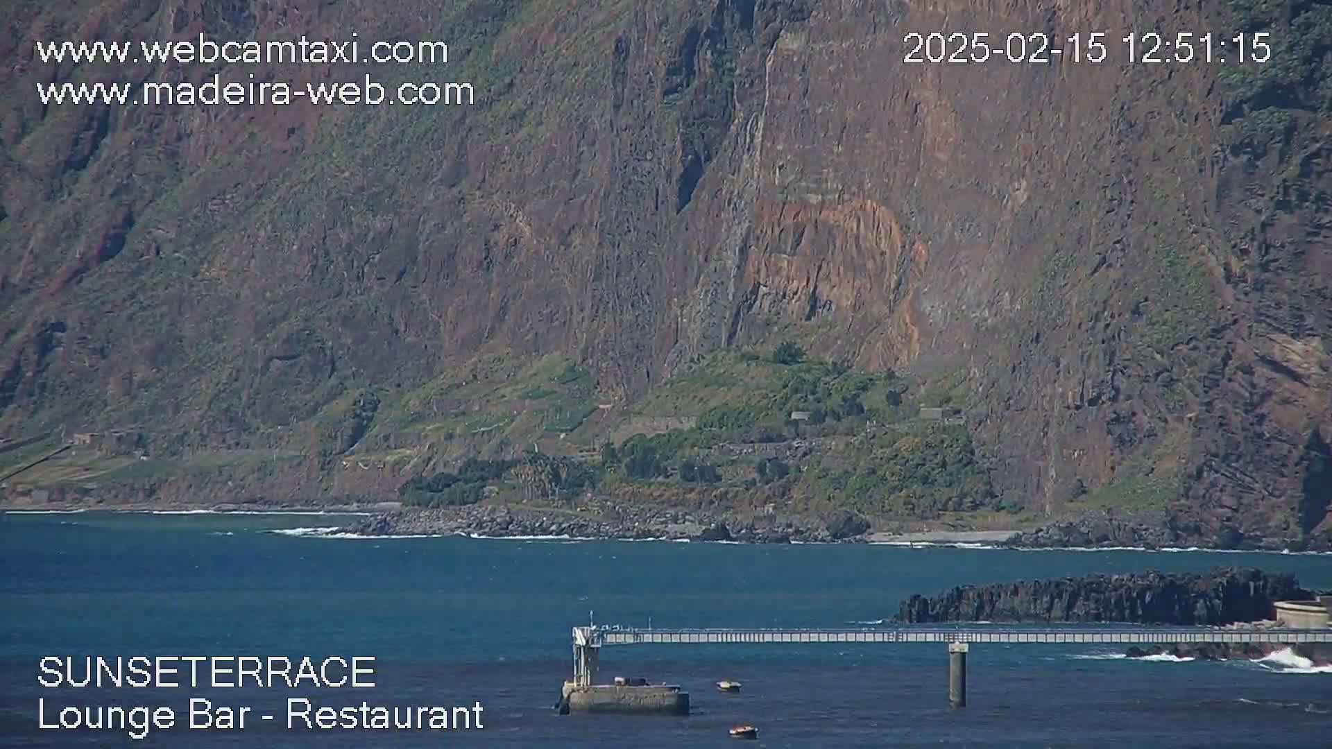 A long pier extends over a calm ocean toward a rocky shore and a large, dark cliff face under a sunny sky.