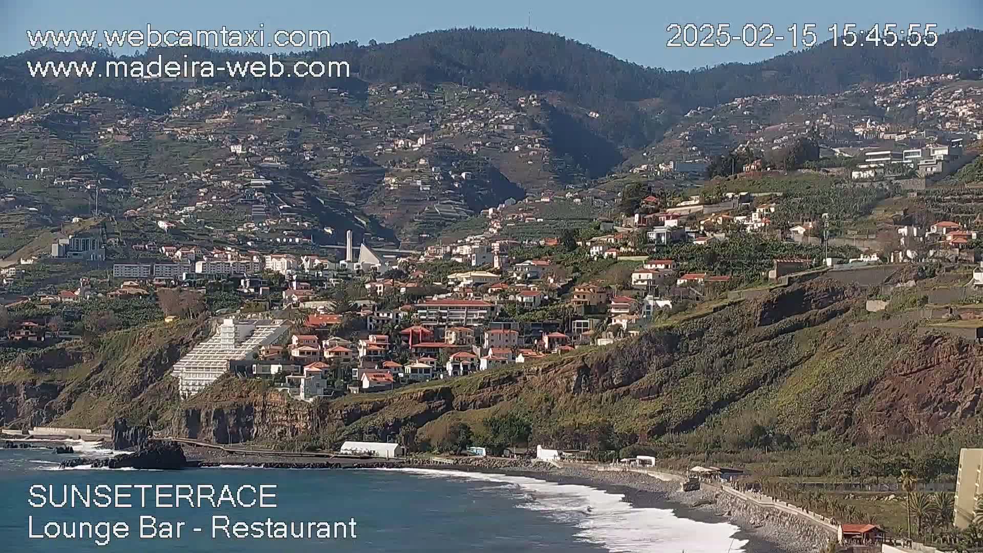 A coastal town nestled on a hillside overlooking a dark-sand beach with gentle waves under a clear, sunny sky.