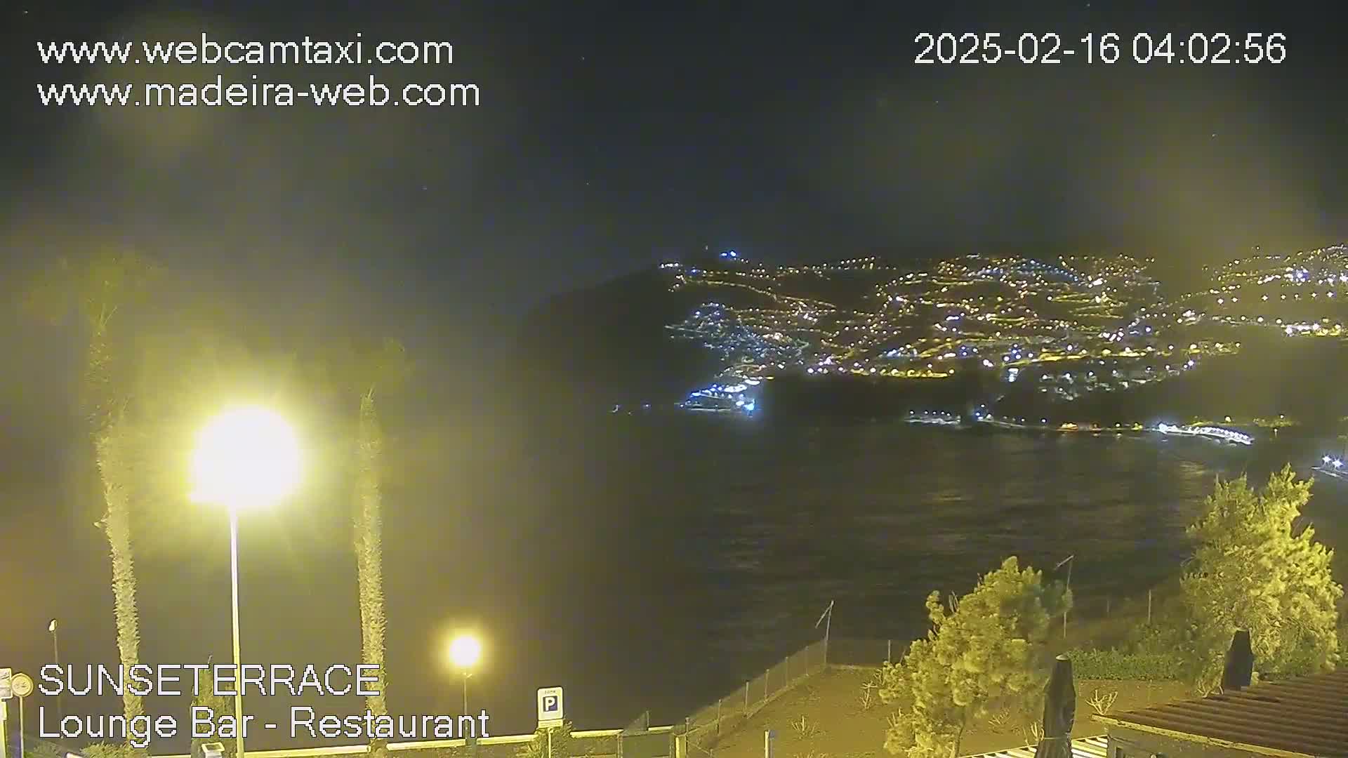A nighttime view of a brightly lit coastal town across a dark body of water, seen from a higher vantage point under a clear sky.