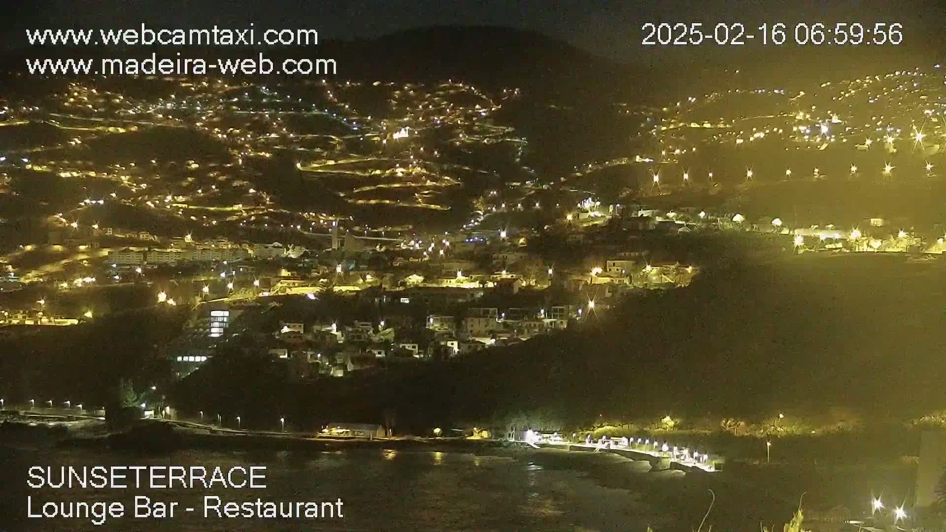 A nighttime view of a hillside town illuminated by lights, extending down to a dark body of water at the base.