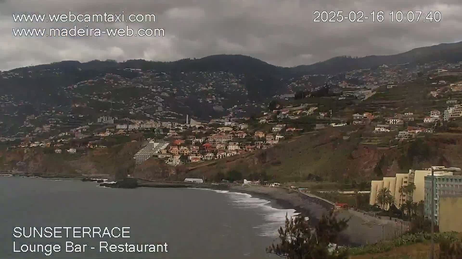 A coastal town nestled on a hillside under an overcast sky overlooks a dark-sand beach with gentle waves.