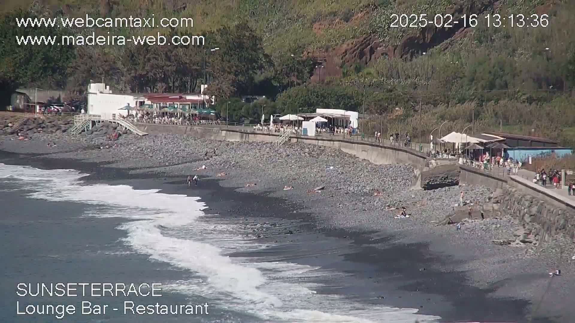 A dark-pebbled beach with people sunbathing and lounging near a beachfront restaurant and bar, with waves lapping the shore on a sunny day.