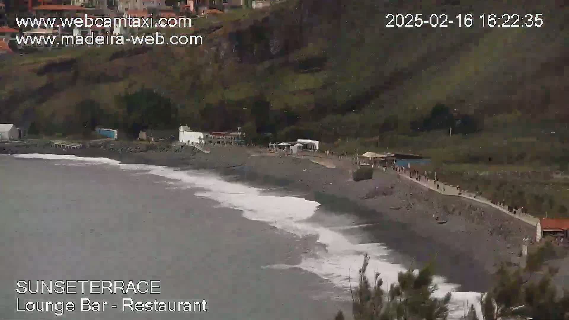 A dark-grey pebble beach with people walking along the shore,  waves breaking on the beach, and buildings behind it, in overcast conditions.