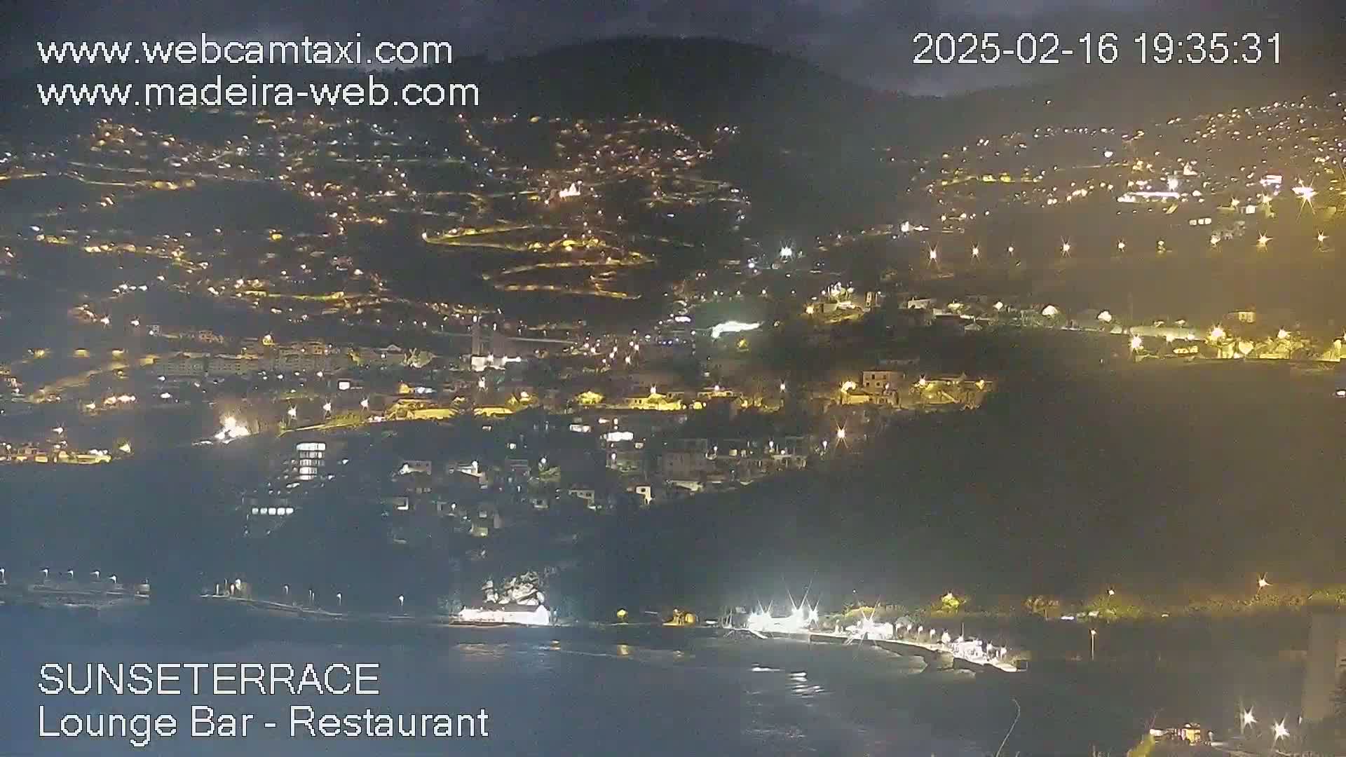 A nighttime view of a hillside town illuminated by many lights, overlooking a dark body of water under a clear sky.
