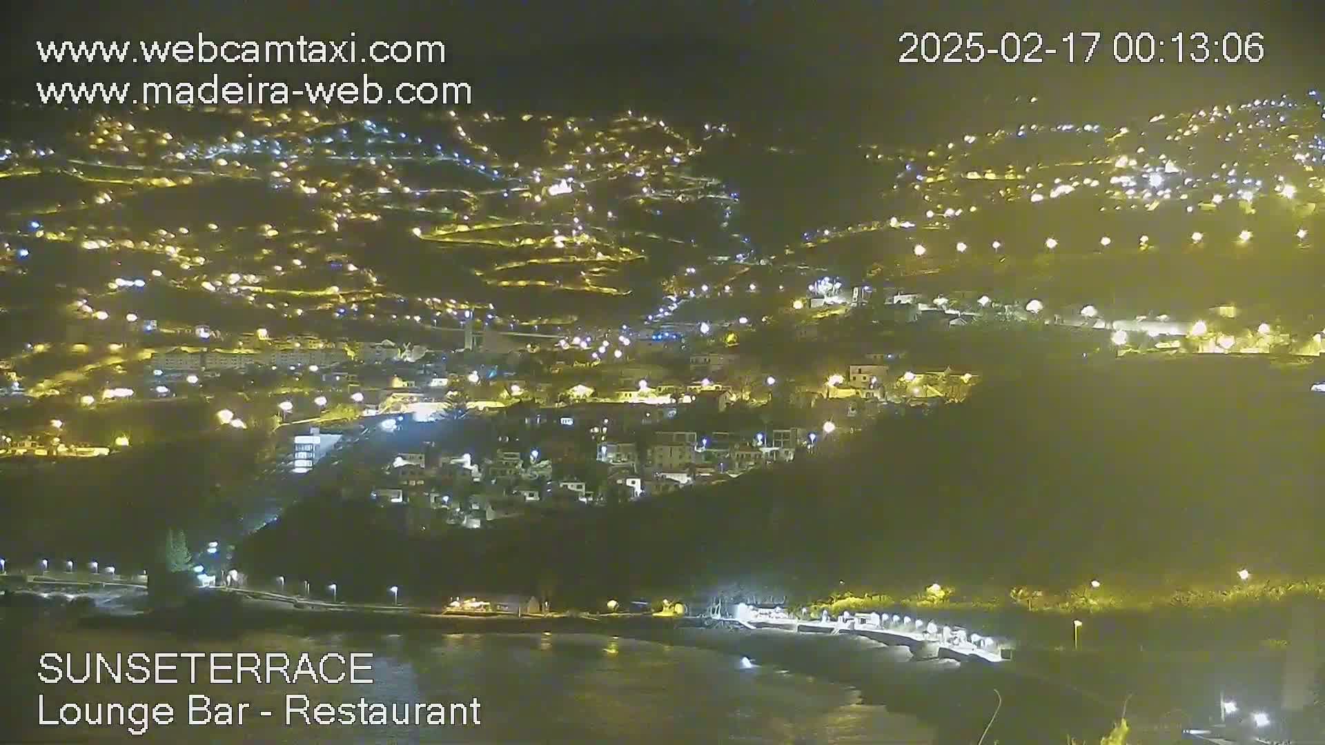 A nighttime, long shot of a coastal town illuminated by many lights, situated on a hillside overlooking a dark body of water.