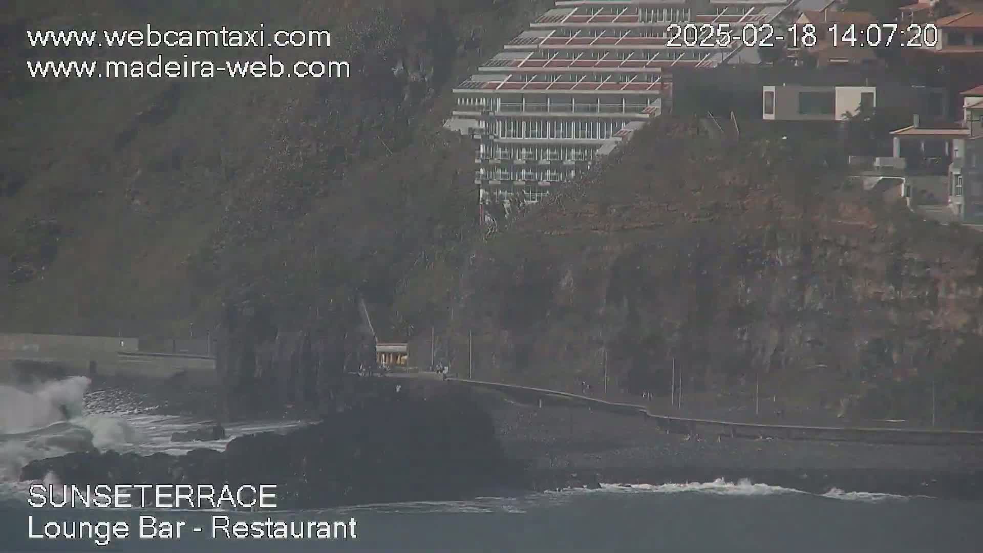 A rocky coastline with a multi-story building perched on a cliff overlooks a dark-colored beach with moderate waves crashing against the shore.