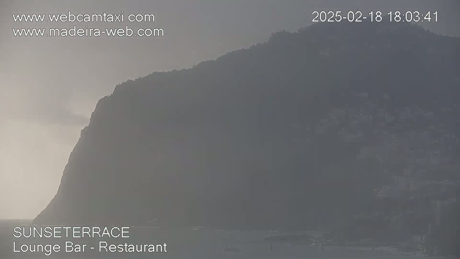A hazy view shows a large, dark hill or mountain overlooking a body of water under a muted, overcast sky.