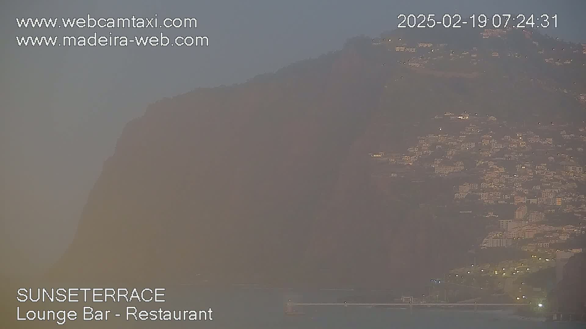 A hazy twilight view shows a hillside town nestled against a large, dark mountain.