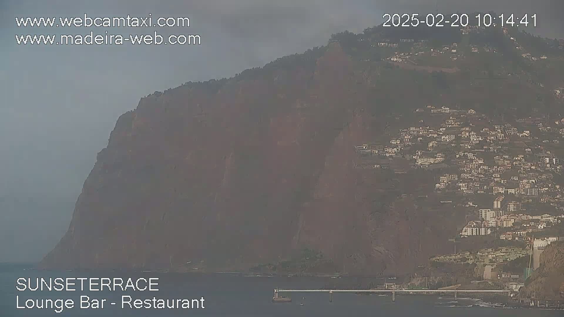 A hazy view shows a large, dark cliff overlooking a town and the ocean.