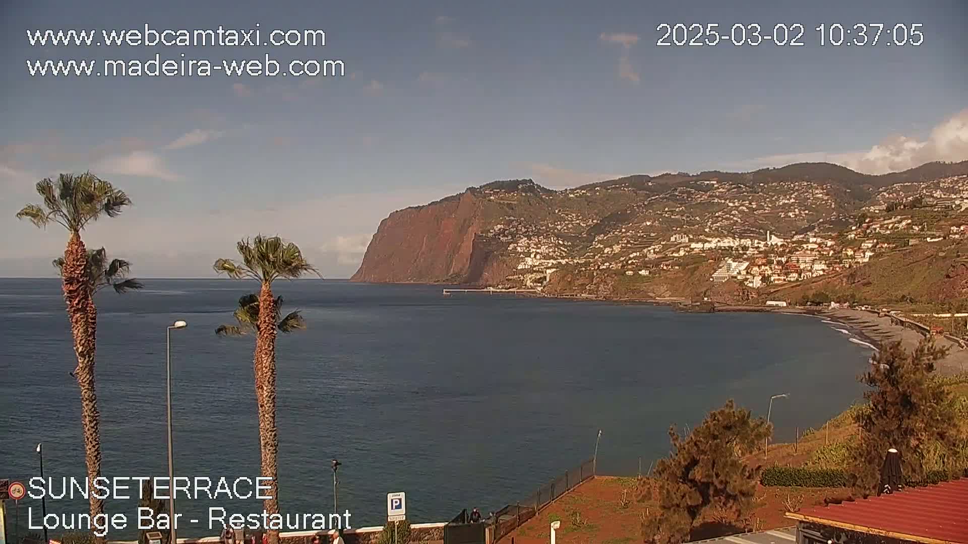 A calm ocean scene with a dark-colored beach, a coastal town built into a hillside, and a large reddish-brown cliff in the background under a mostly clear sky.