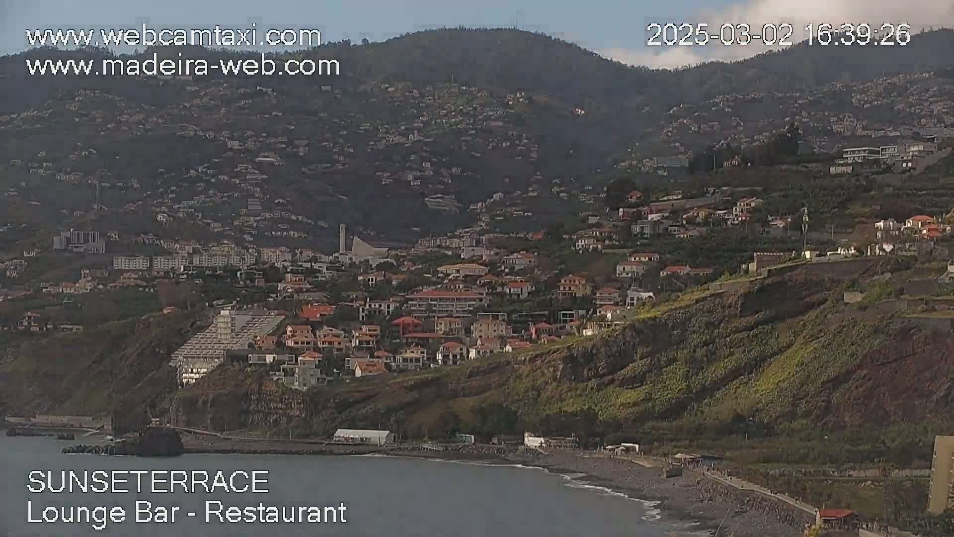 A coastal town nestled on a hillside overlooking a calm ocean under a mostly clear sky.