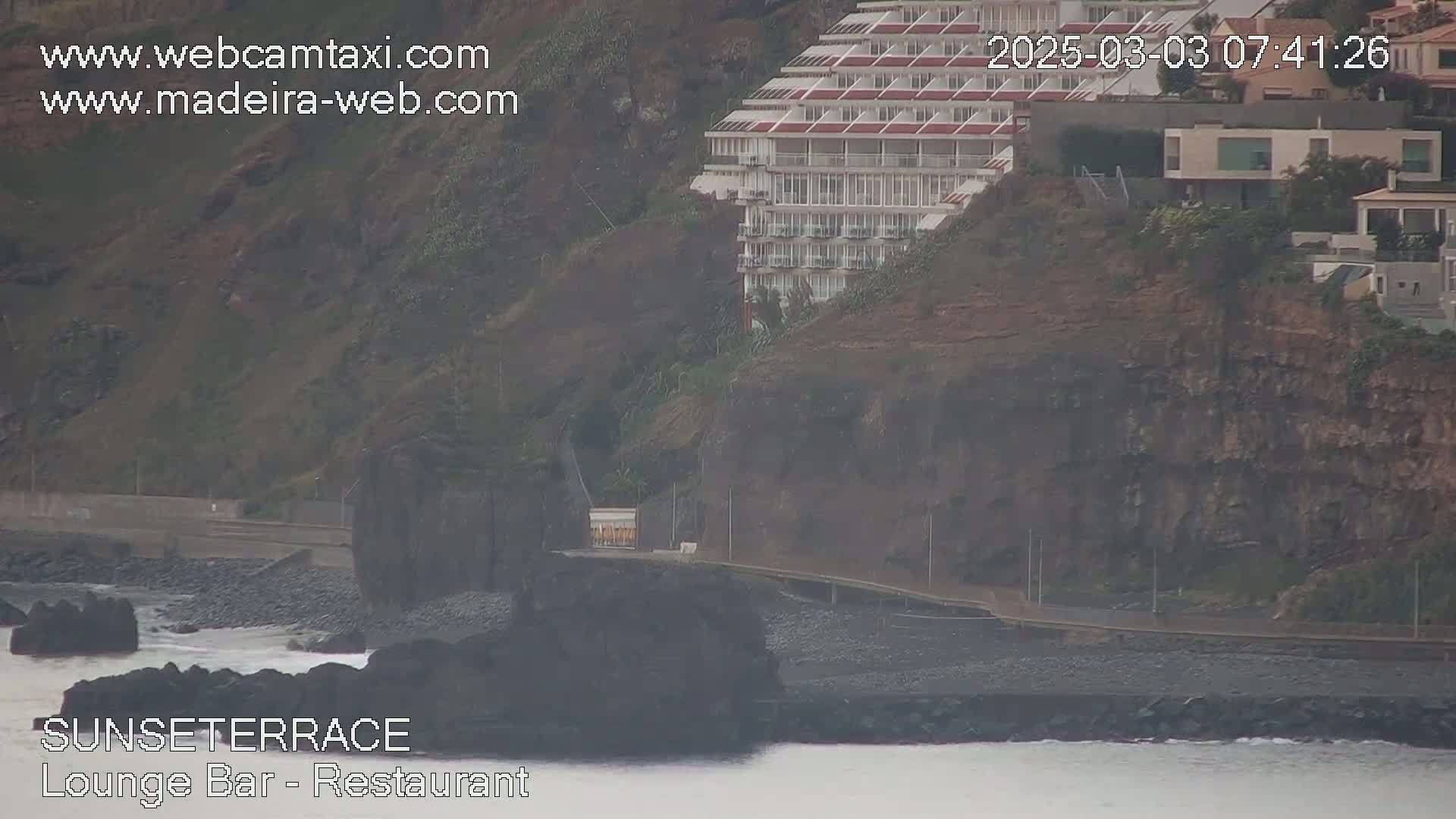 A coastal scene shows a multi-story building perched on a rocky cliff overlooking a dark, pebbly beach with a calm ocean and hazy weather conditions.