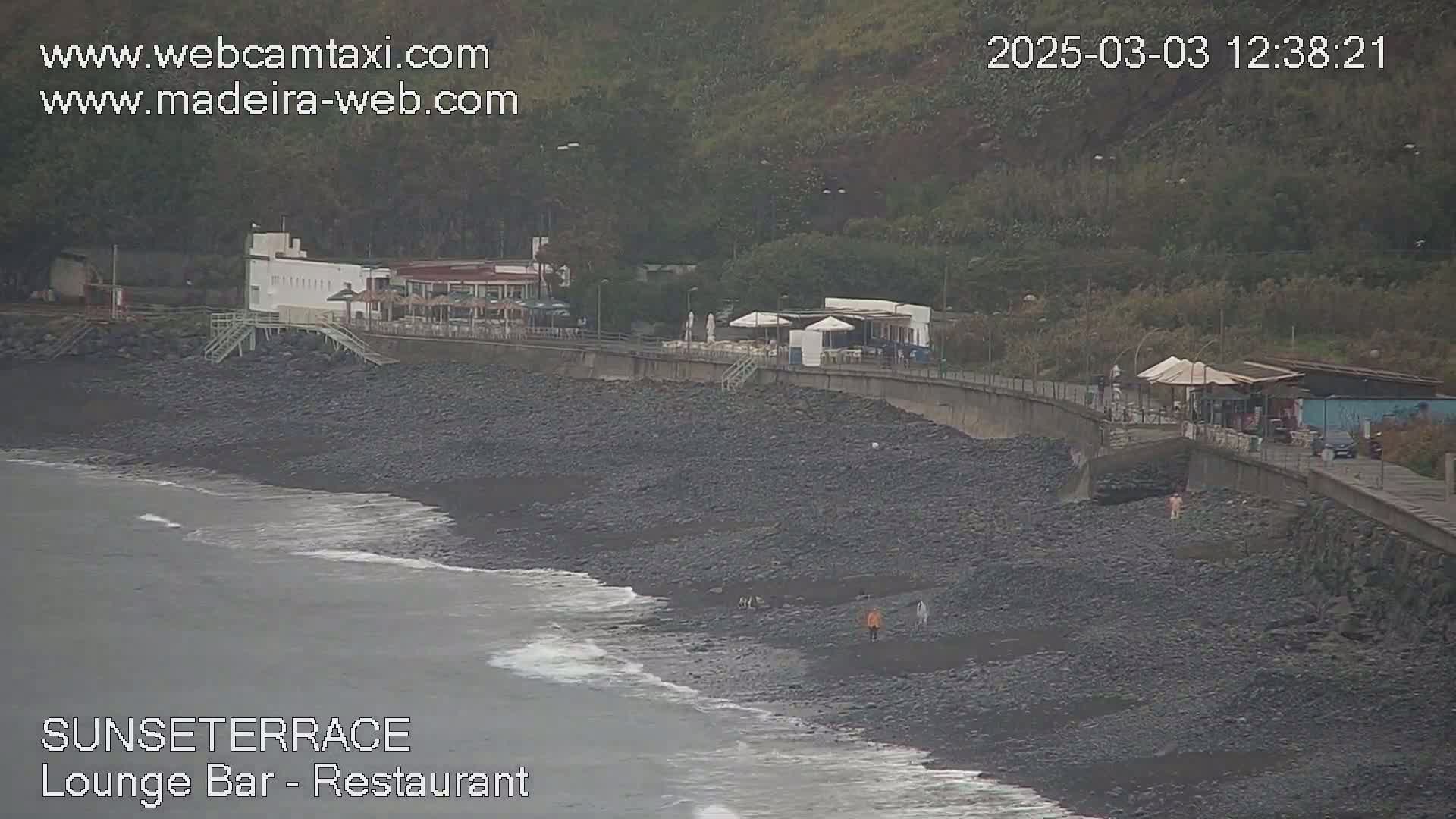 A dark-colored, rocky beach with a low wall separating it from buildings and a paved walkway, all under an overcast sky.