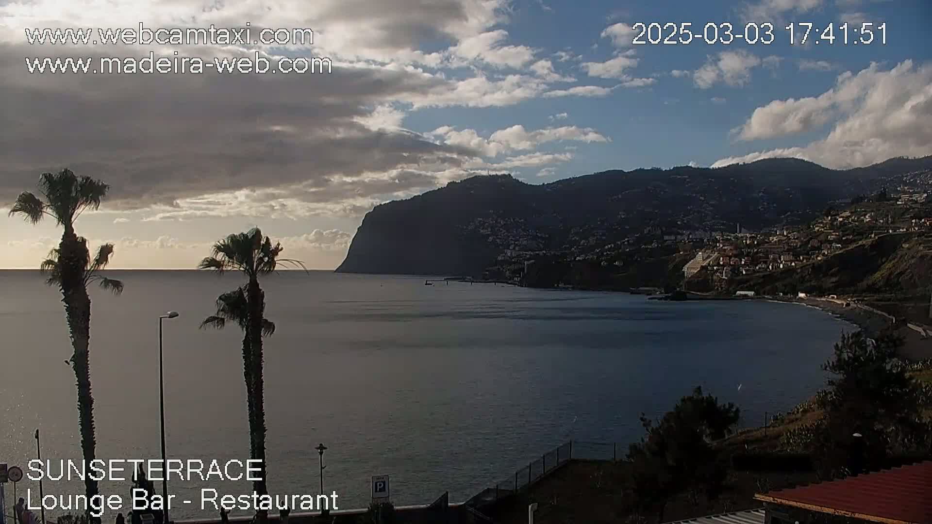 A partly cloudy sunset over a calm ocean bay, with palm trees in the foreground and a mountainside town in the background.