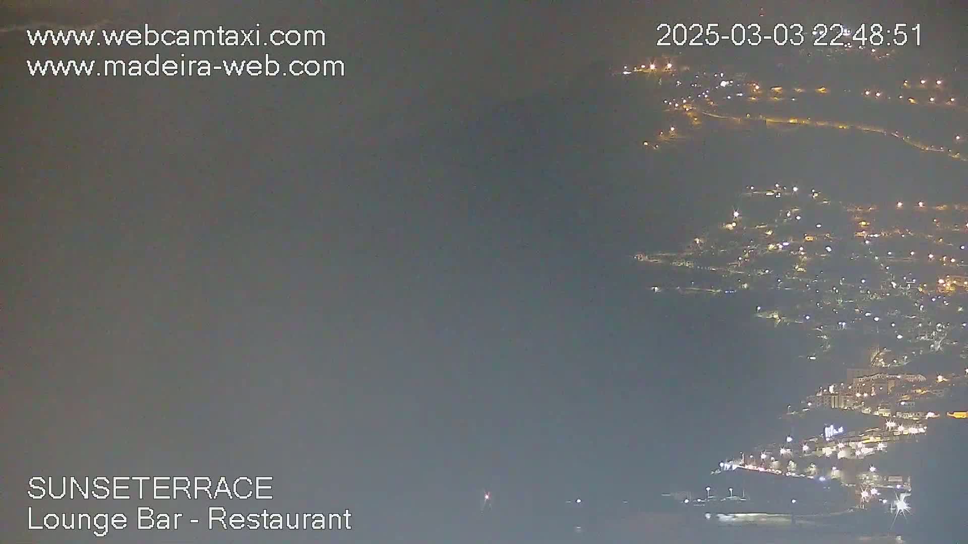 A nighttime long shot shows a coastal town with many lights illuminated against a hazy, dark sky.