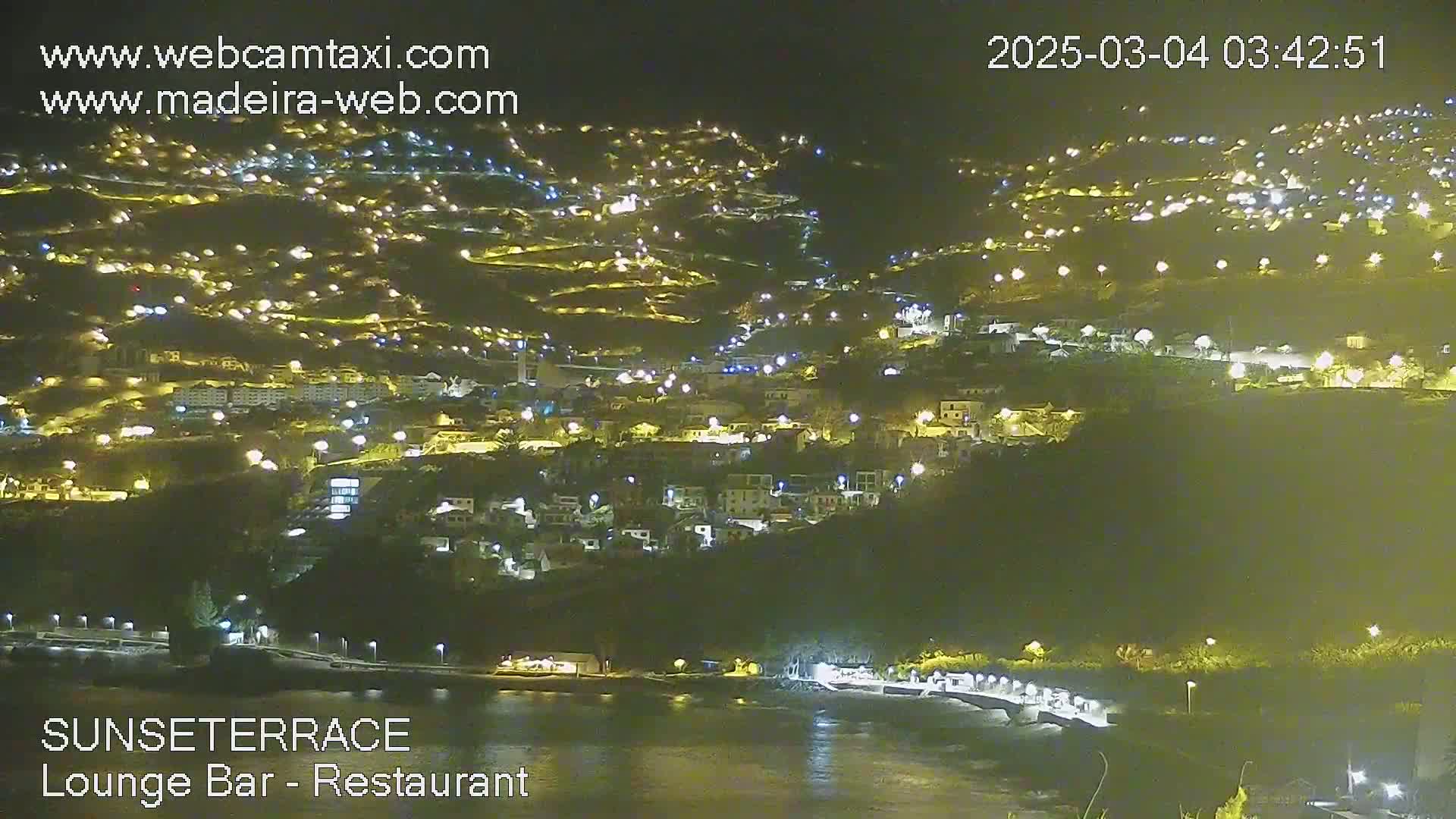 A nighttime, coastal town illuminated by numerous lights climbs a hillside that slopes down to a dark body of water.