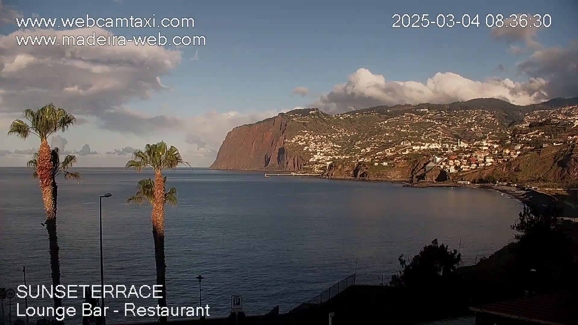A coastal town nestled against a large, reddish cliff face is viewed from across a calm ocean under a partly cloudy sky.
