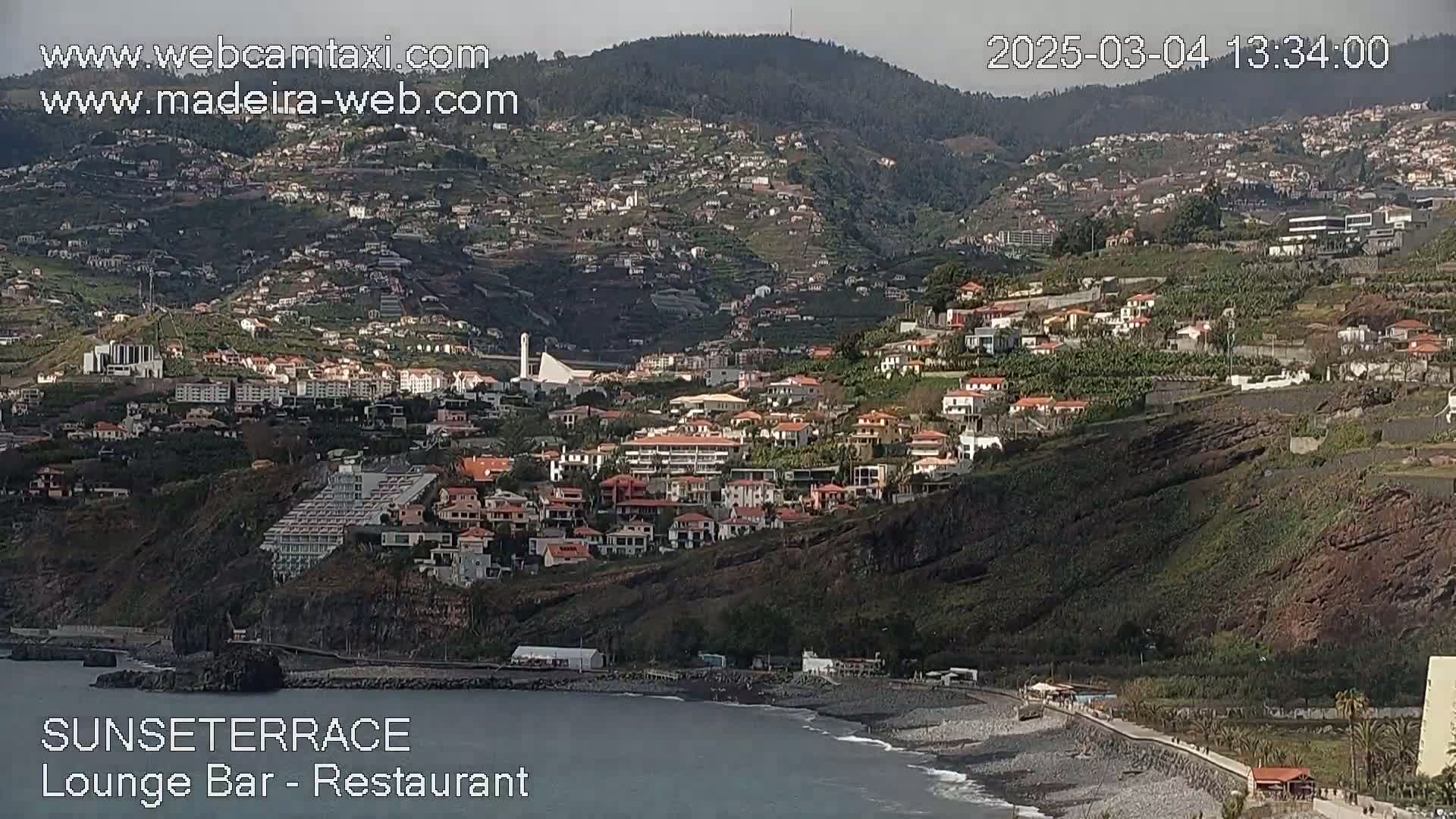 A coastal town nestled on a hillside overlooking a dark-colored beach and ocean under a mostly cloudy sky.