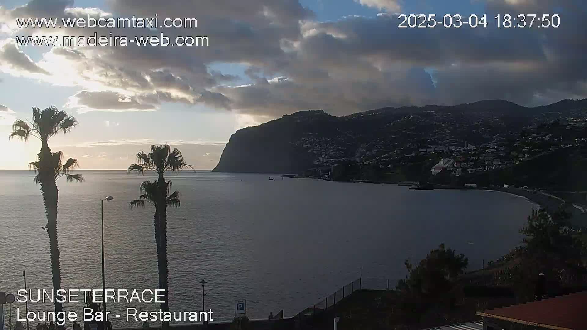 A partly cloudy sunset over a calm ocean with a coastal town and a large cliff in the background, viewed from a vantage point with palm trees in the foreground.