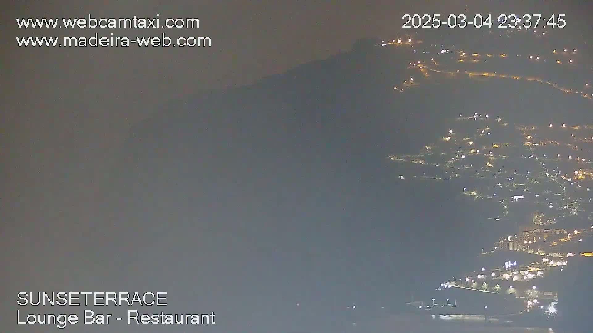 A nighttime long shot shows a hillside town with many lights illuminated against a dark, overcast sky.