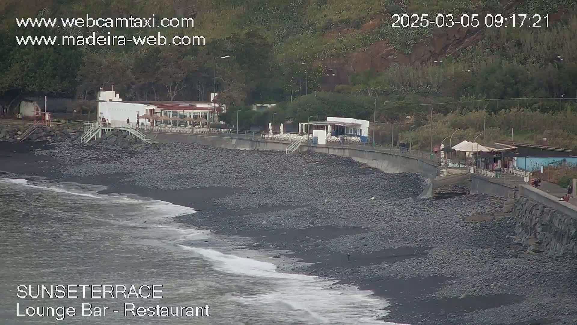A dark pebble beach with gentle waves laps against the shore, next to a restaurant and walkway backed by lush green vegetation on a hillside.