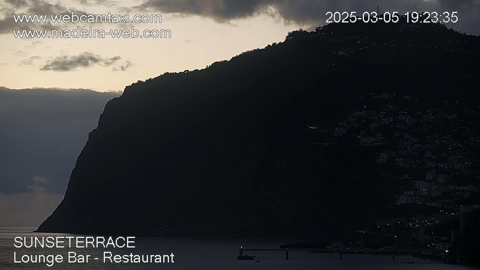 A large, dark mountain silhouetted against a twilight sky overlooks a dark coastline dotted with faint lights.