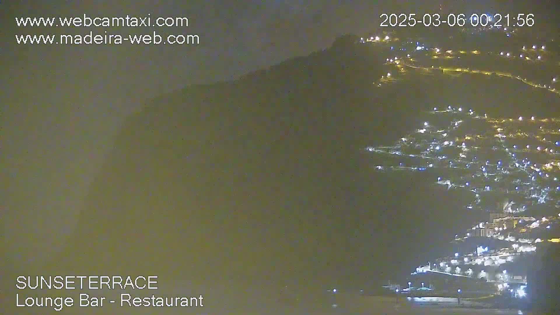 A nighttime long shot shows a hillside town illuminated by many small lights, under a dark, cloudy sky.