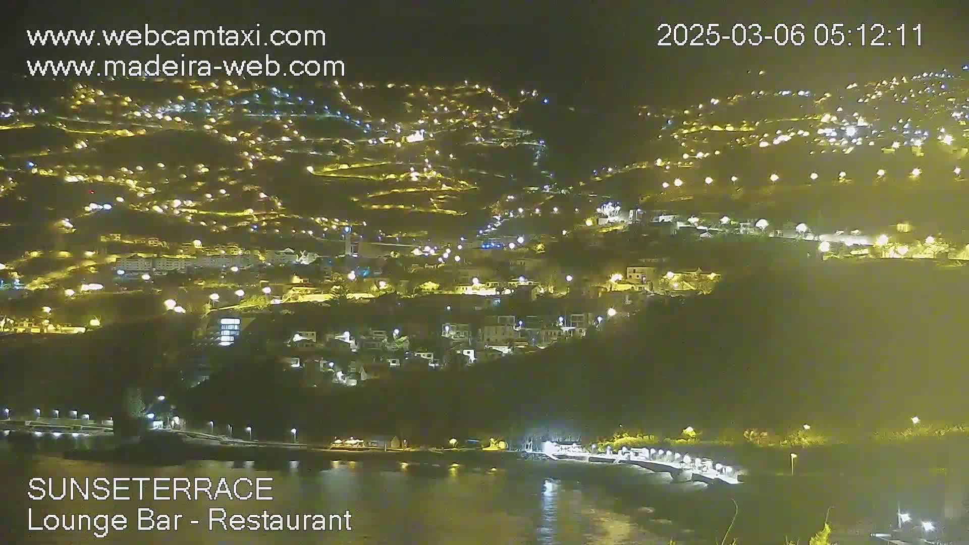 A nighttime view of a hillside town illuminated by many lights, next to a dark body of water.