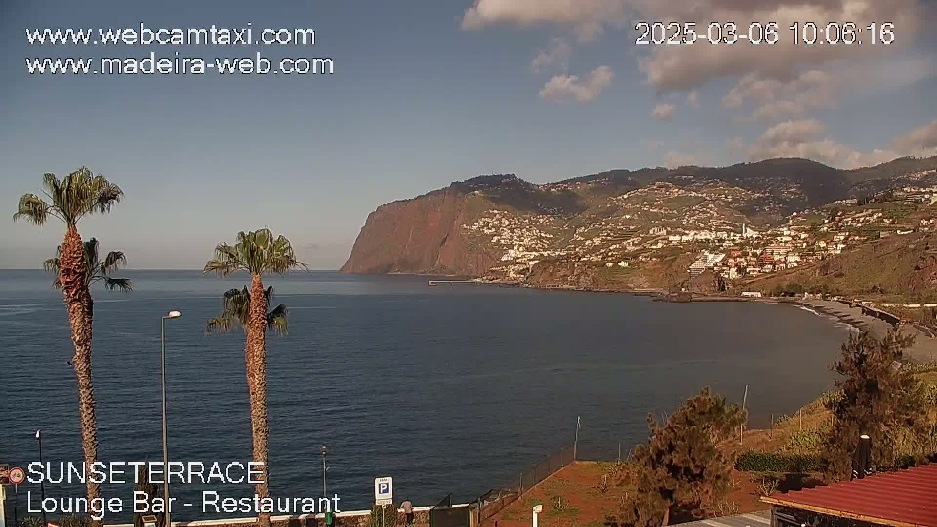 A partly cloudy day overlooking a calm ocean with a coastal town and a high cliff in the distance, two palm trees in the foreground.