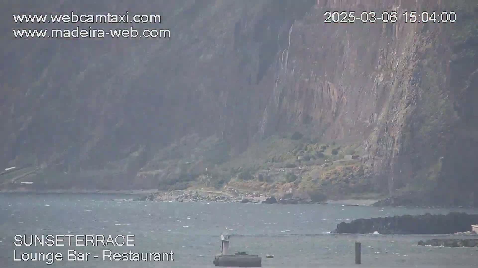 A rocky cliff overlooks a choppy ocean with a small structure in the water.