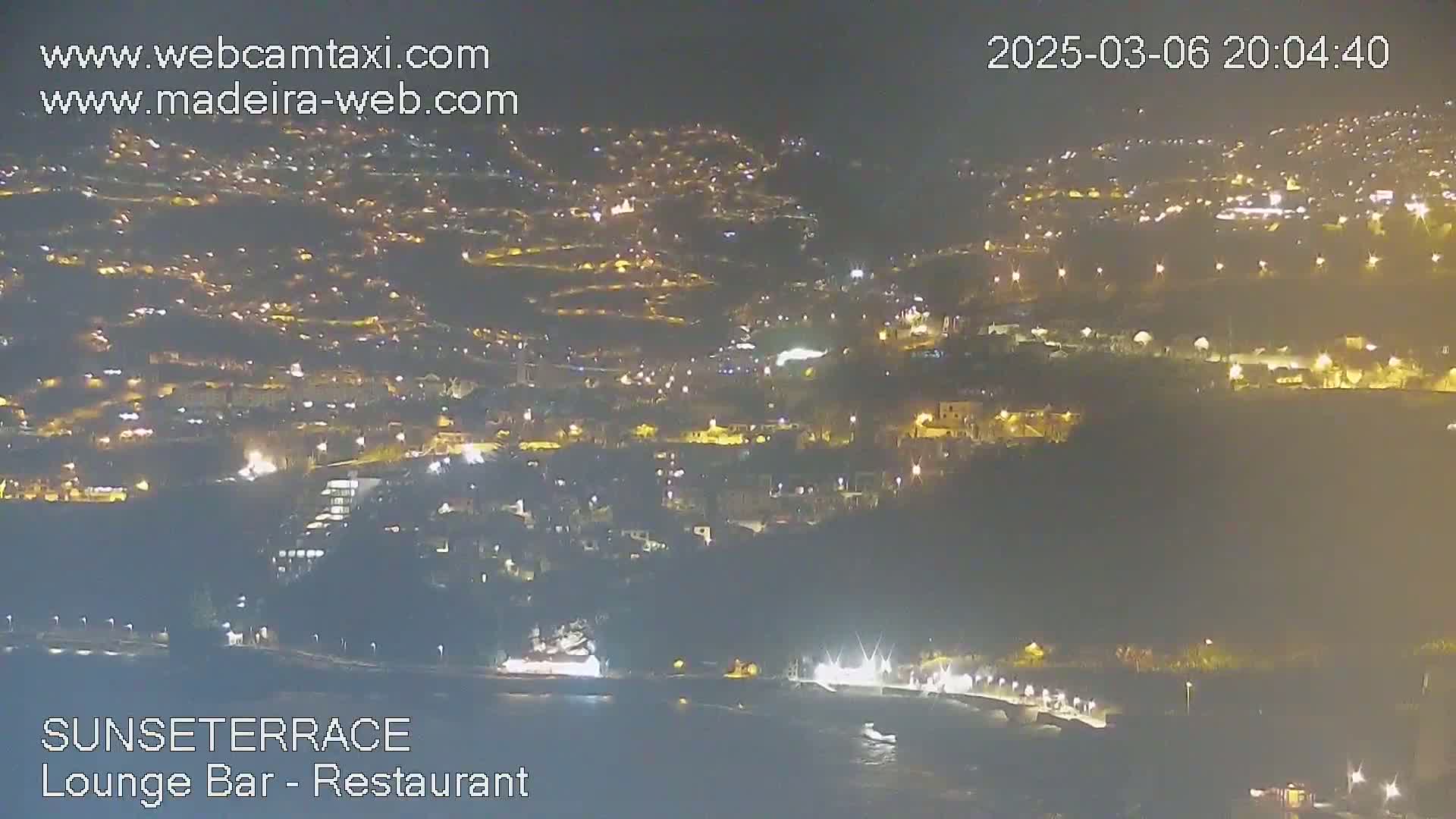 A nighttime, high-angle view shows a coastal town illuminated with many lights, extending down to a dark body of water.
