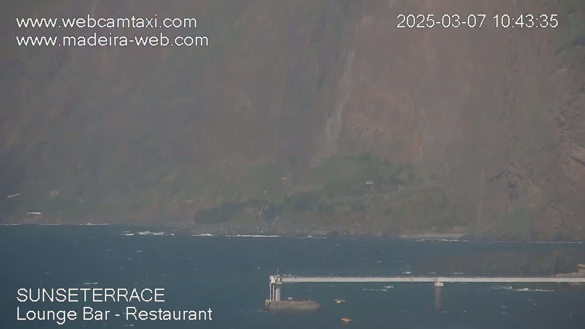 A long, low pier extends into a somewhat choppy ocean, with hazy brown hills rising in the background.