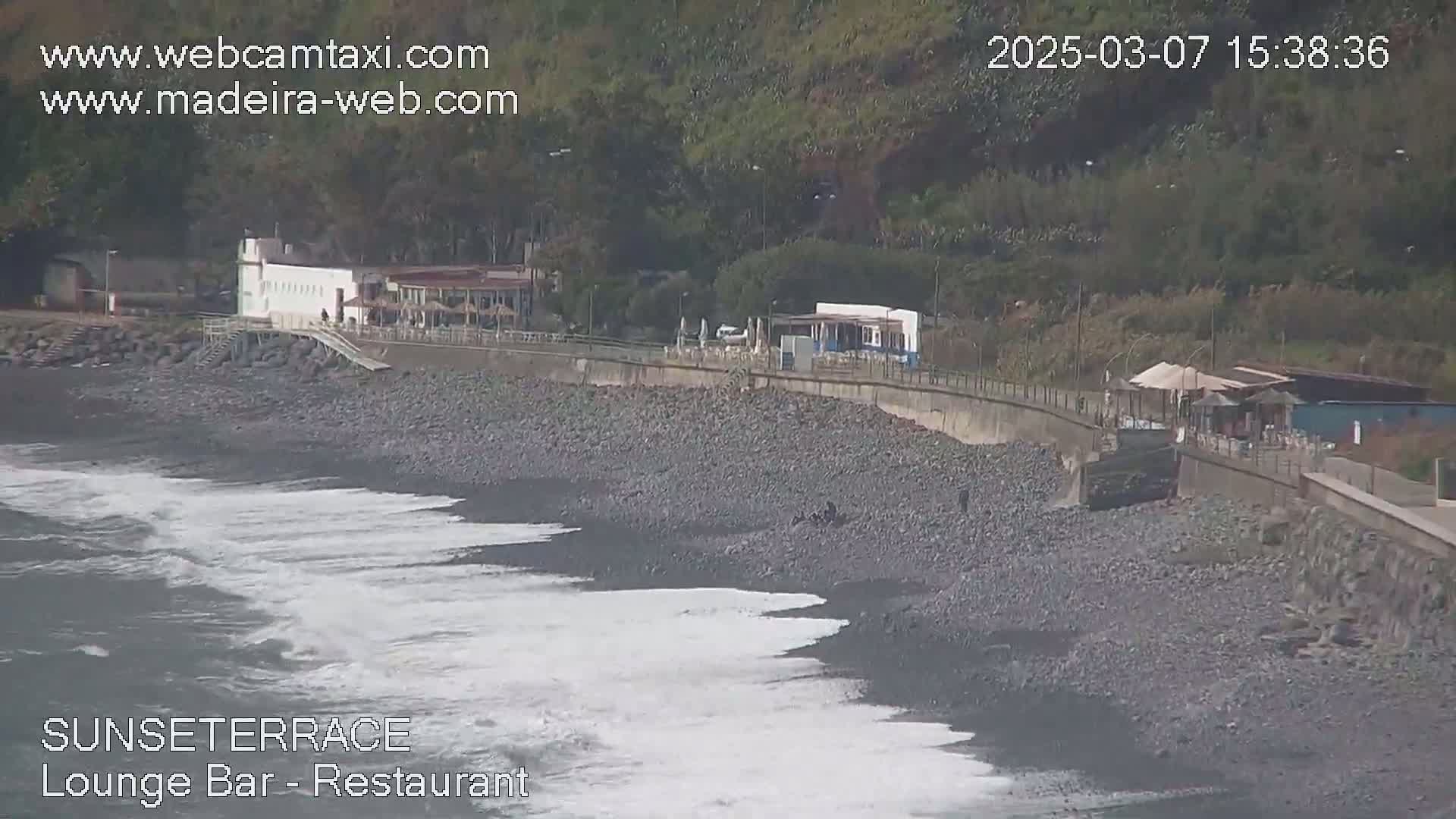 A rocky beach with small waves laps against the shore, next to a restaurant and a seawall backed by green hills.